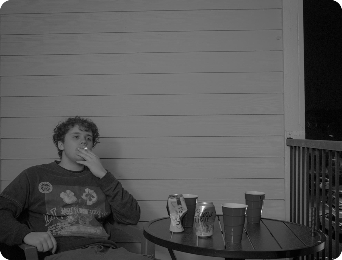 black and white image of a college student smoking a cigarette with empty beer cans and solo cups on a table next to him