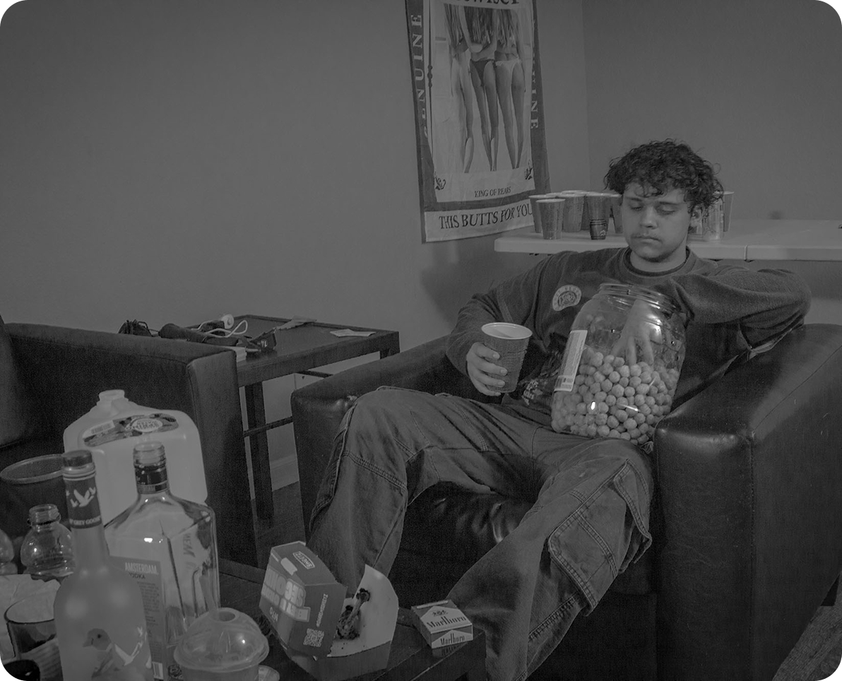 black and white image of a college kid sticking his hand into a bin of cheese balls in front of a messy table with alcohol, cigarettes, and food on it